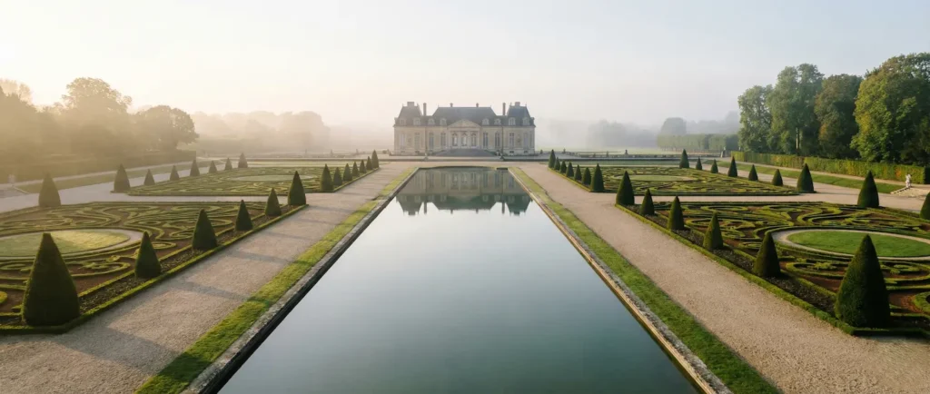 Vista panorámica 21:9 de parterres de boj simétricos y estanque reflectante en jardín francés.