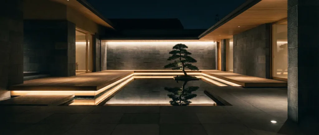 Vista panorámica 21:9 de patio japonés con iluminación nocturna y materiales nobles.