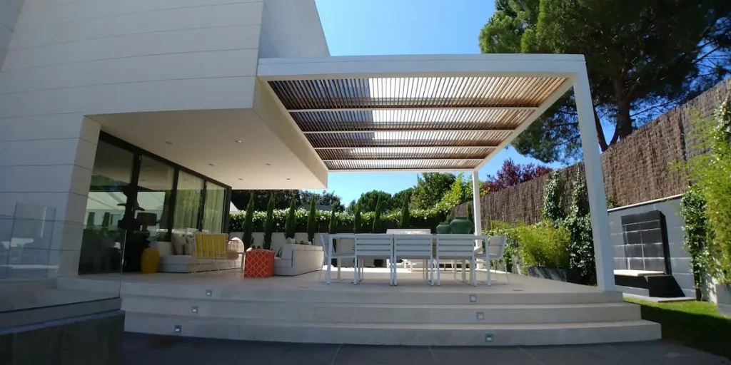 Vista interior de una pérgola bioclimática adosada de aluminio blanco de Proyectos Xanadú, con lamas móviles en el techo sobre una zona de sofás de exterior frondosa.