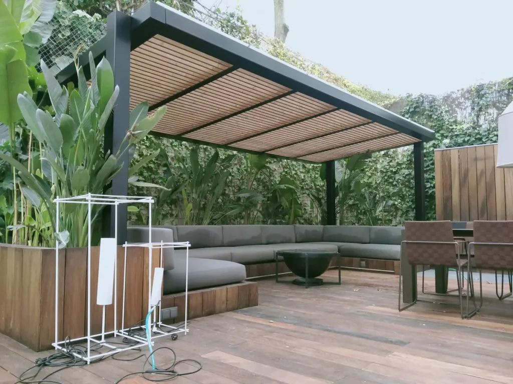 Elevada terraza residencial con pérgola de aluminio oscuro adosada, techo de cristal fijo, suelo de deck técnico imitación madera clara rampa y jardineras a juego con vegetación por Proyectos Xanadú.