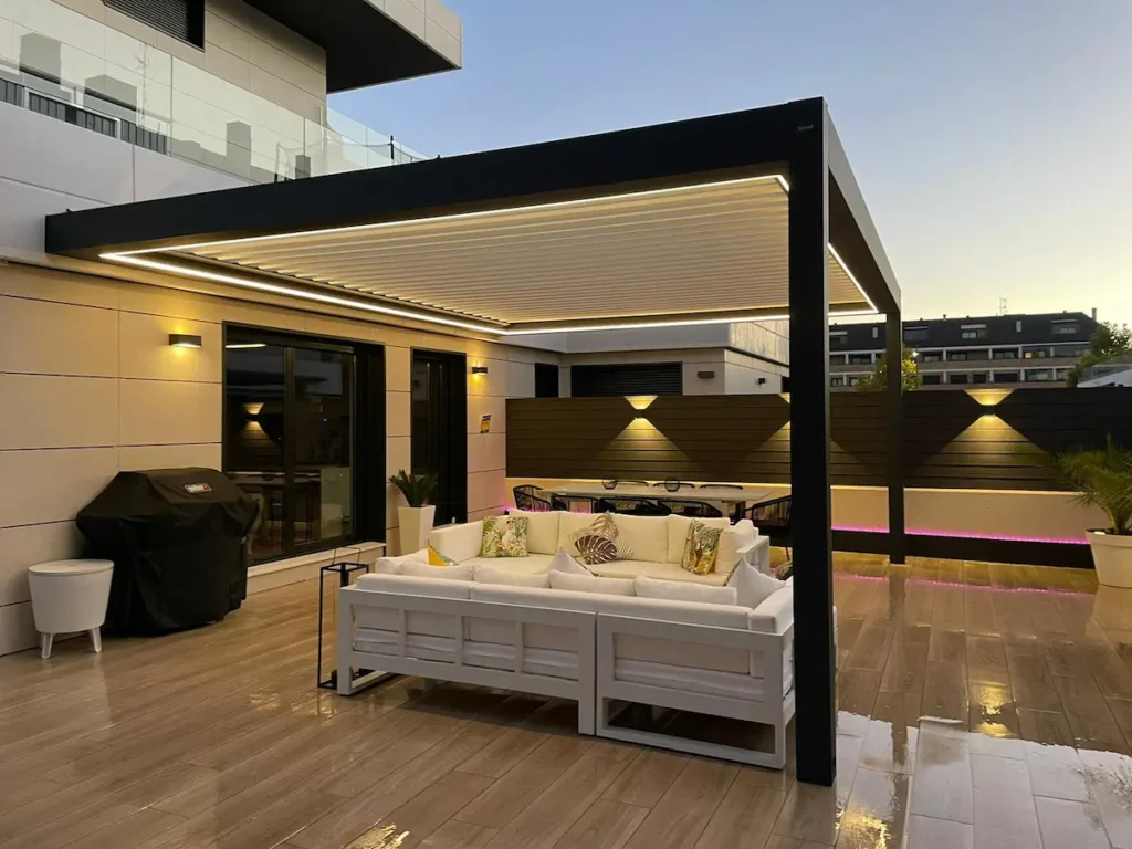 Detalle de una estructura de aluminio oscuro con techo de lona blanca tensada de Proyectos Xanadú adosada a una fachada con sombras de árboles.