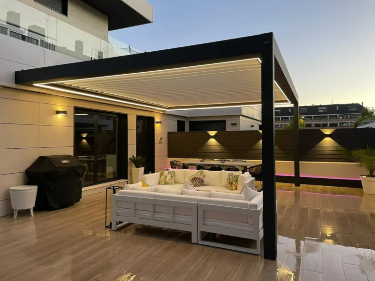 Detalle de una estructura de aluminio oscuro con techo de lona blanca tensada de Proyectos Xanadú adosada a una fachada con sombras de árboles.